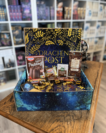 Das Produktbild zeigt vorn eine geöffnete Drachenpost in blauem Schuppen-Design. Man sieht in der Box ein in ein dunkellilafarbenes Drachenpost-Poster verpacktes Buch und zum Thema Schokolade passende Süßigkeiten. Im Hintergrund sieht man die Bücherregale des Verlagsladens.